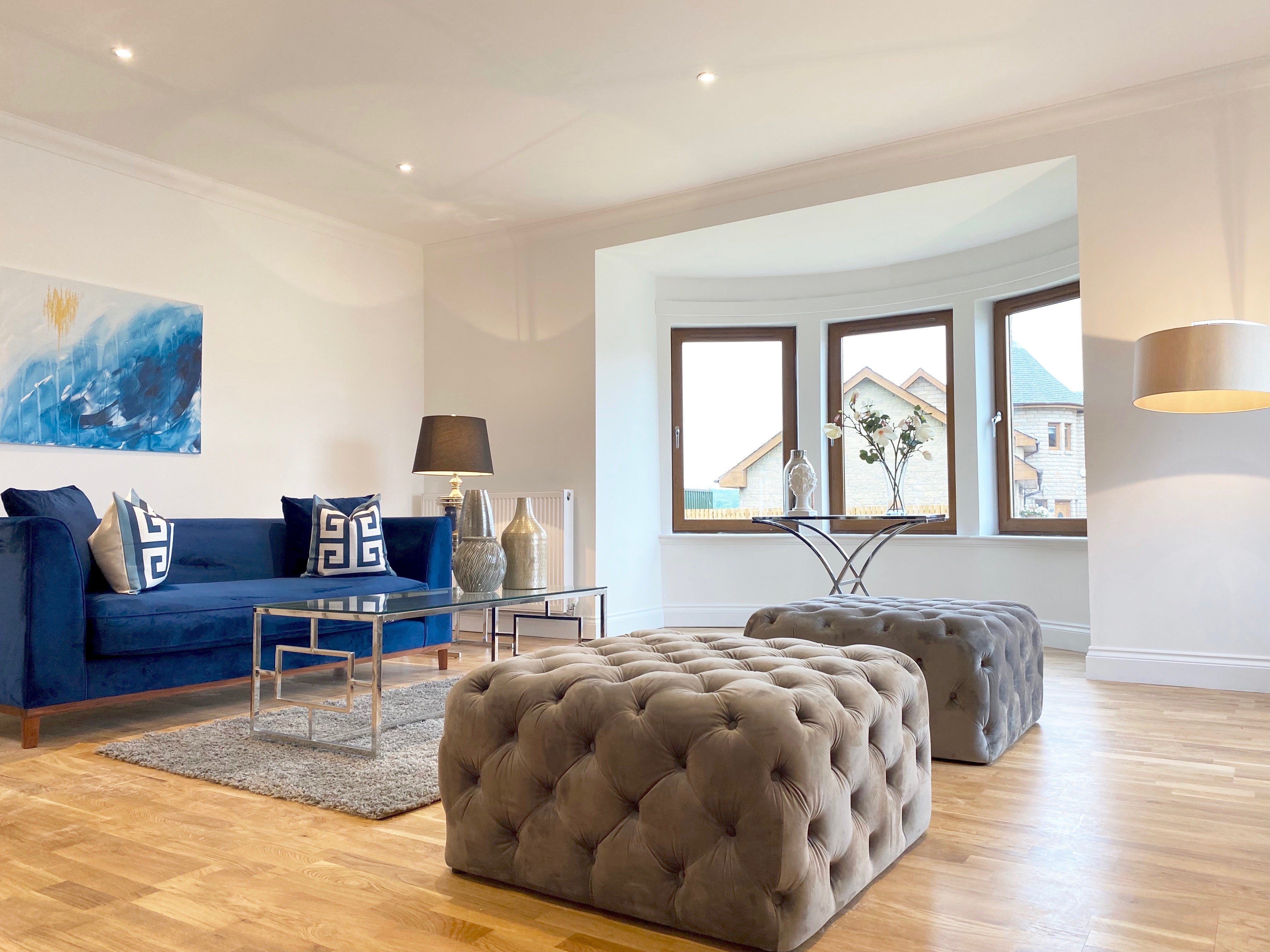 Royal blue velvet sofa, stainless steel coffee table with glass top, two grey ottomans, bright blue artwork, floor lamp, table lamp, grey rug